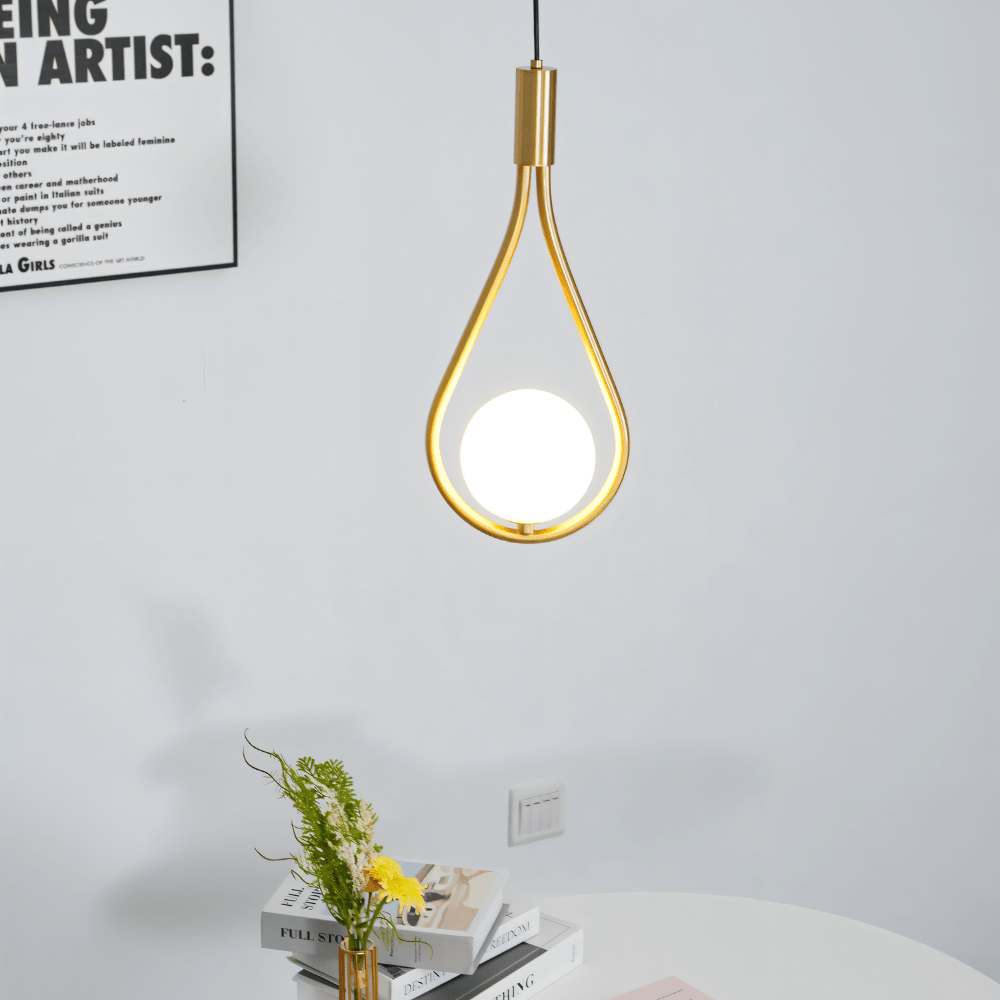 Clérachic brass teardrop pendant with an opal globe hanging against a pale wall above a round table with books and flowers.