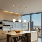 Row of three Clérachic brass teardrop pendants with opal globes over a marble kitchen island with black stools and a city view.