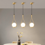 Three Clérachic brass teardrop pendants with glowing opal globes lined up over a dining table with a vase and a bowl.