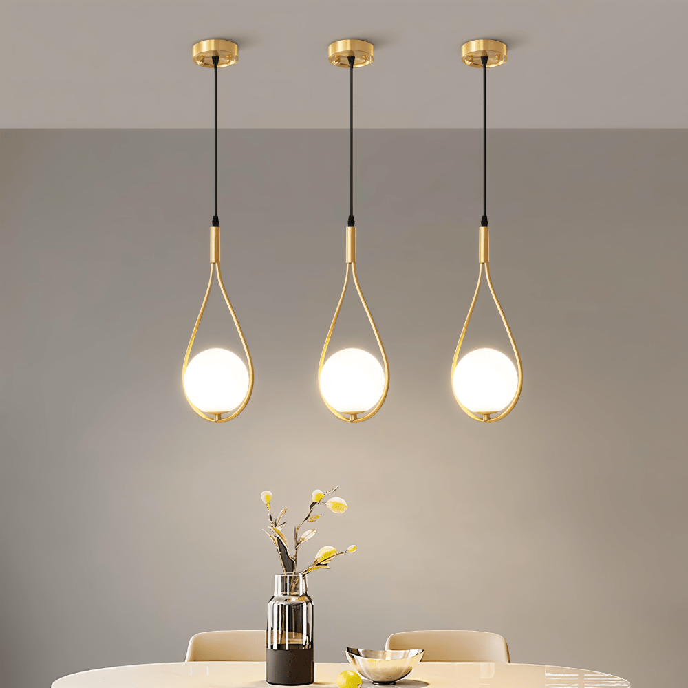 Three Clérachic brass teardrop pendants with glowing opal globes lined up over a dining table with a vase and a bowl.