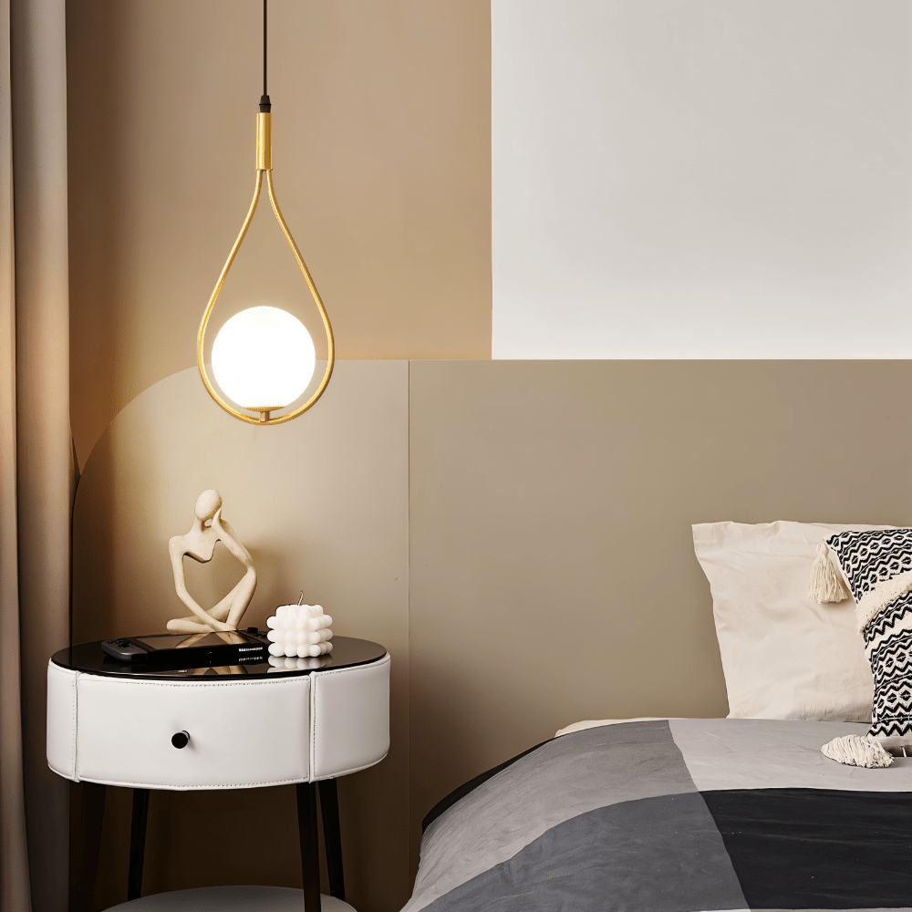 Clérachic brass teardrop pendant with a lit opal globe beside a white leather bedside table, bed and taupe panelled wall.
