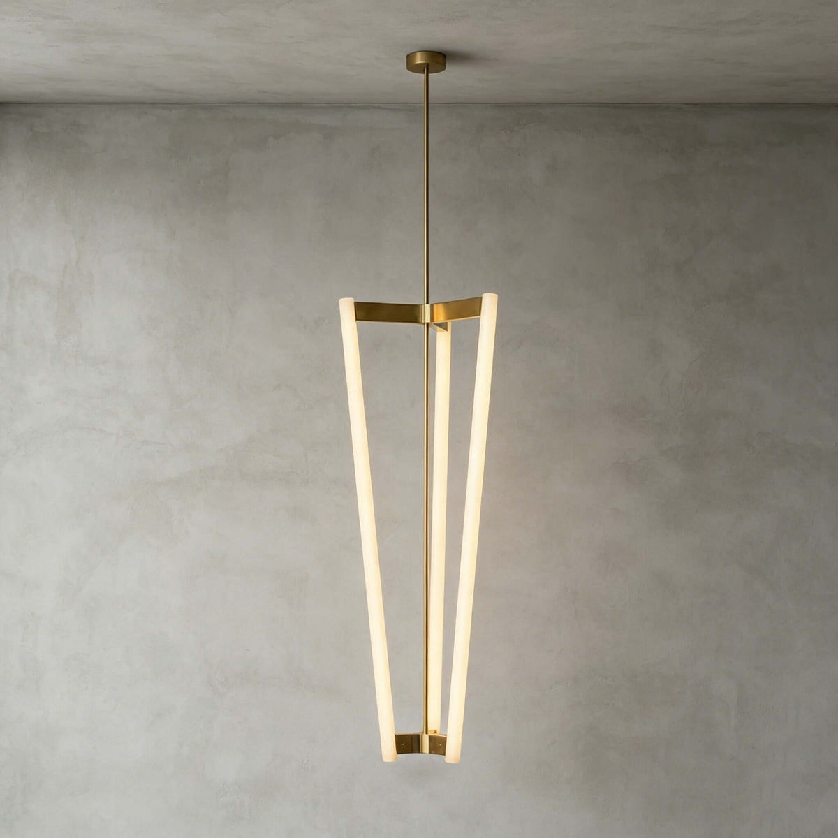 Against a grey plaster wall, this Clérachic pendant hangs from a ceiling canopy and brass rod, three slim LED tubes lit.