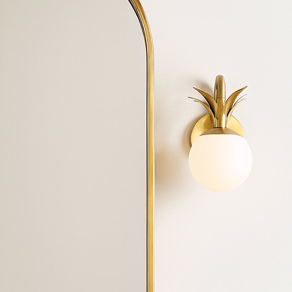 Single Clérachic wall light with one opal glass globe and a brass pineapple crown, beside an arched mirror on a warm neutral wall.
