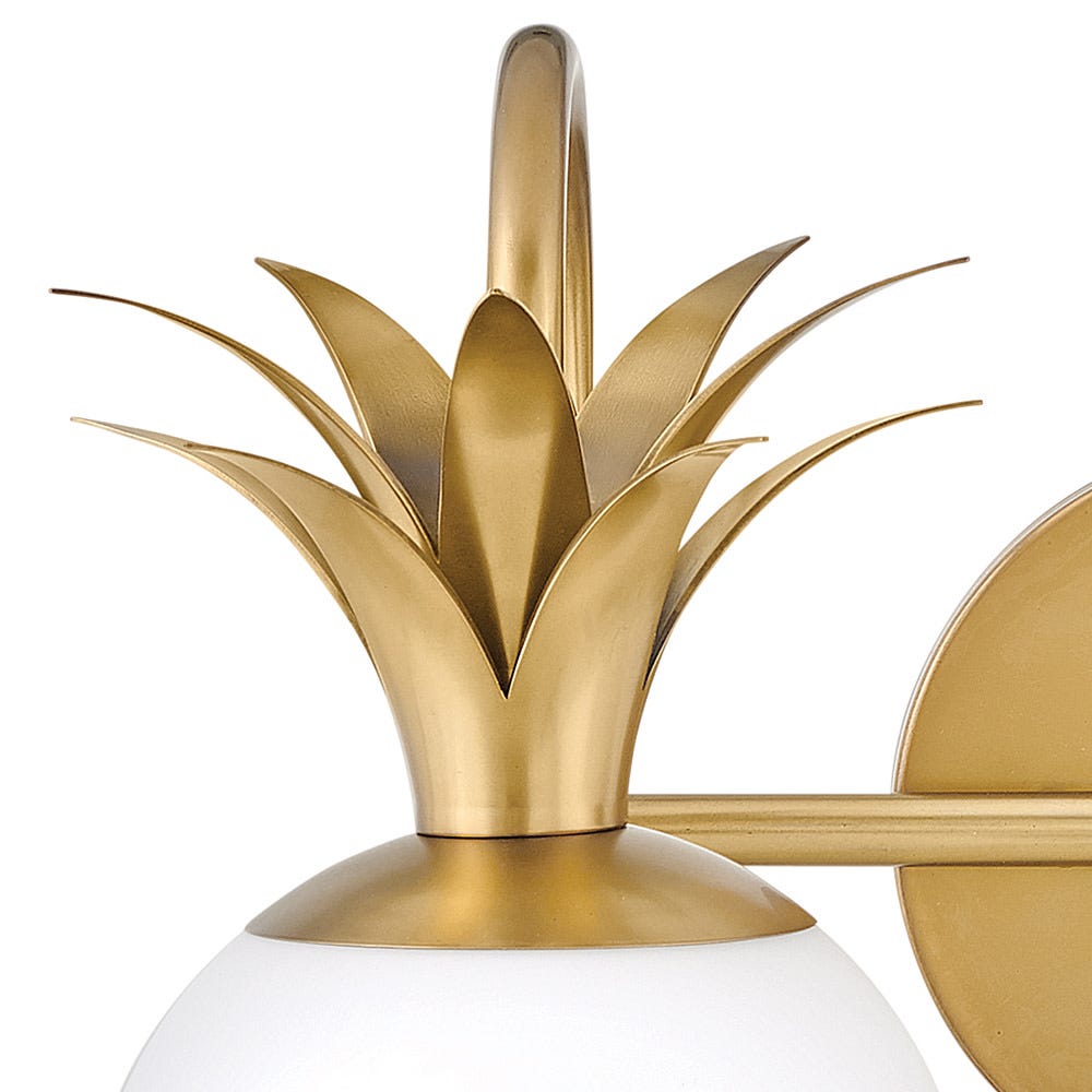 Close view of the brass pineapple-leaf crown and curved arm on this Clérachic wall light, above a white glass globe.