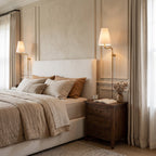 On either side of an upholstered bed, a pair of lit Clérachic wall lights with white fabric shades glow beside linen curtains.