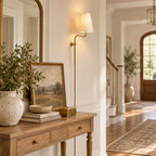 Above a wooden console table with a framed landscape, a lit Clérachic wall light with a brass arm hangs by a hallway.