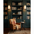 A pair of Clérachic wall lights with brass arms glow on either side of a dark green bookcase above a tan leather armchair.