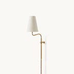 Brass Clérachic wall light with a gently curved arm and a white fabric shade, shown on a pale wall with a twisted cord.