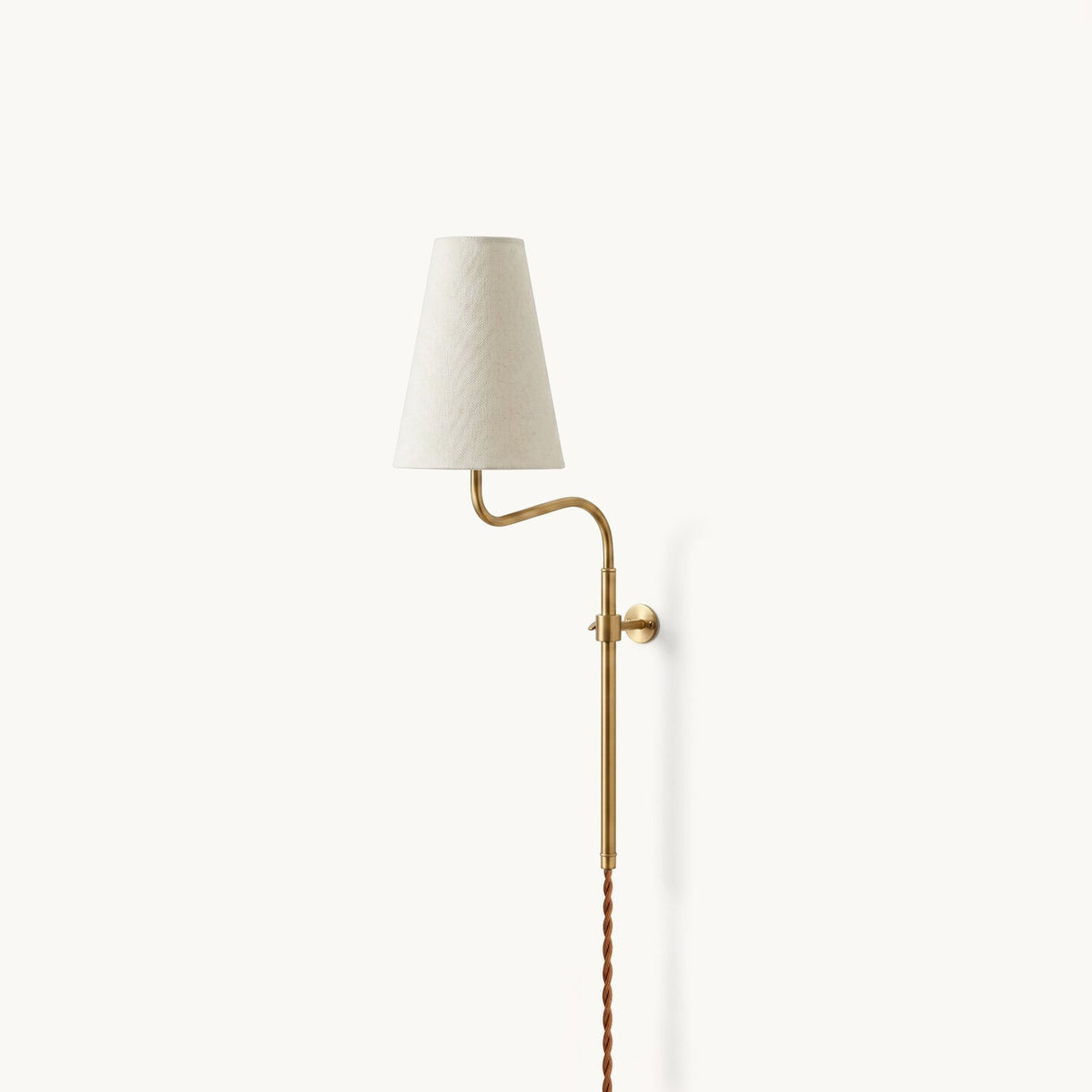 Brass Clérachic wall light with a gently curved arm and a white fabric shade, shown on a pale wall with a twisted cord.