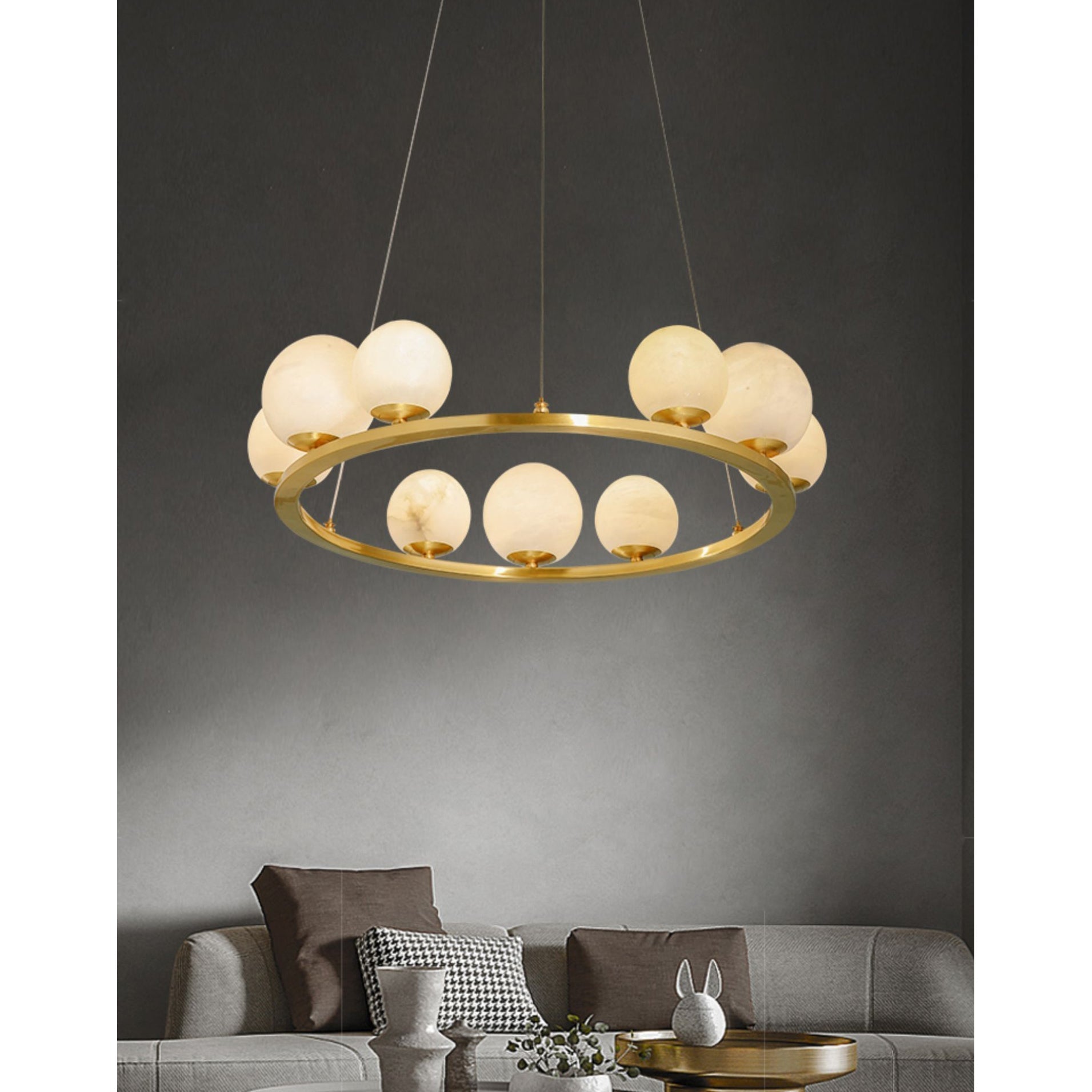 The Clérachic ring chandelier lit above a grey sofa on a dark wall, the alabaster globes reading as a slow cluster of moons.