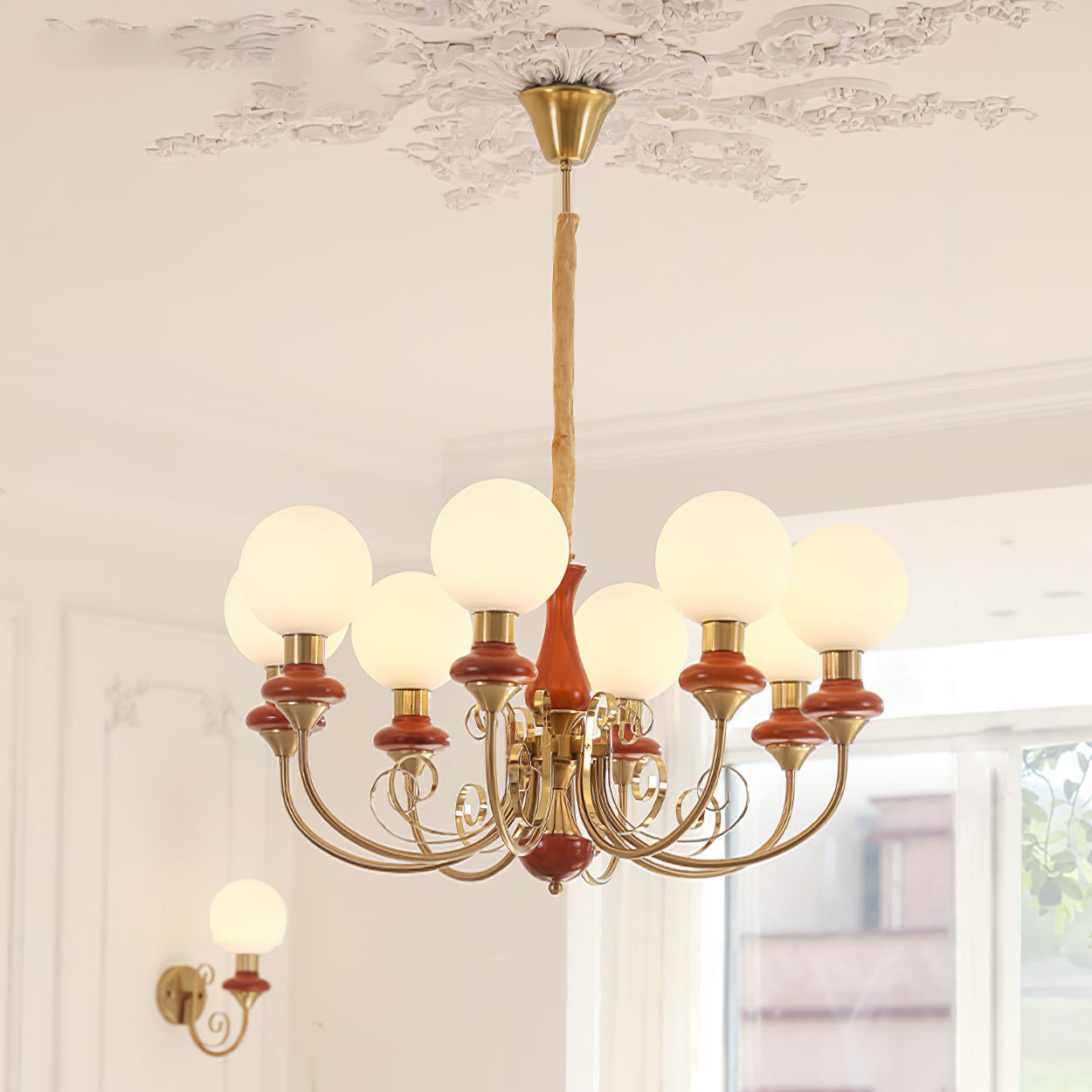 Eight-light Clérachic Brass Scrollwork Chandelier in red-brown, lit above a bright room beside the matching wall light.