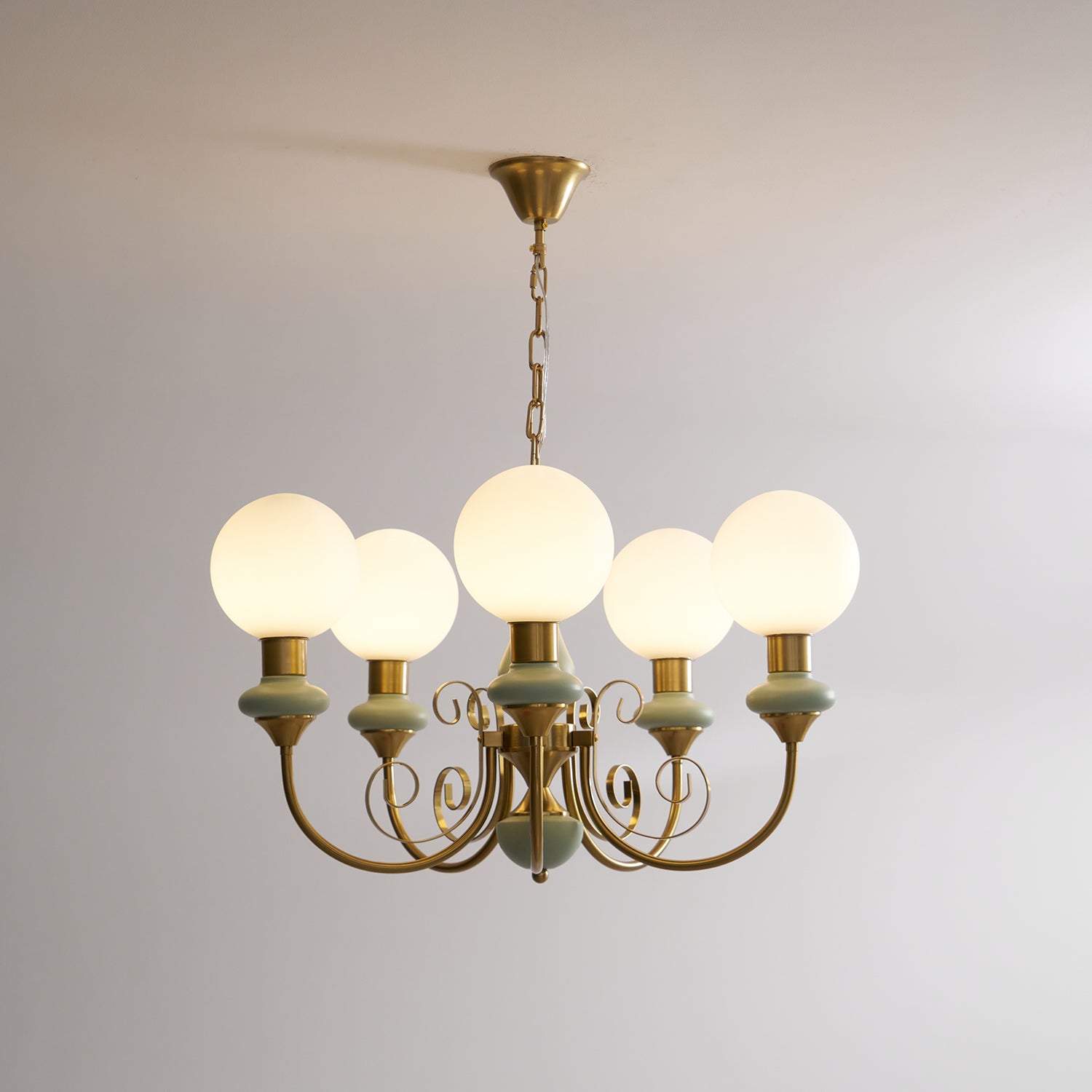 Sage-green Clérachic Brass Scrollwork Chandelier lit on its brass chain, opal globes glowing against a bare wall.
