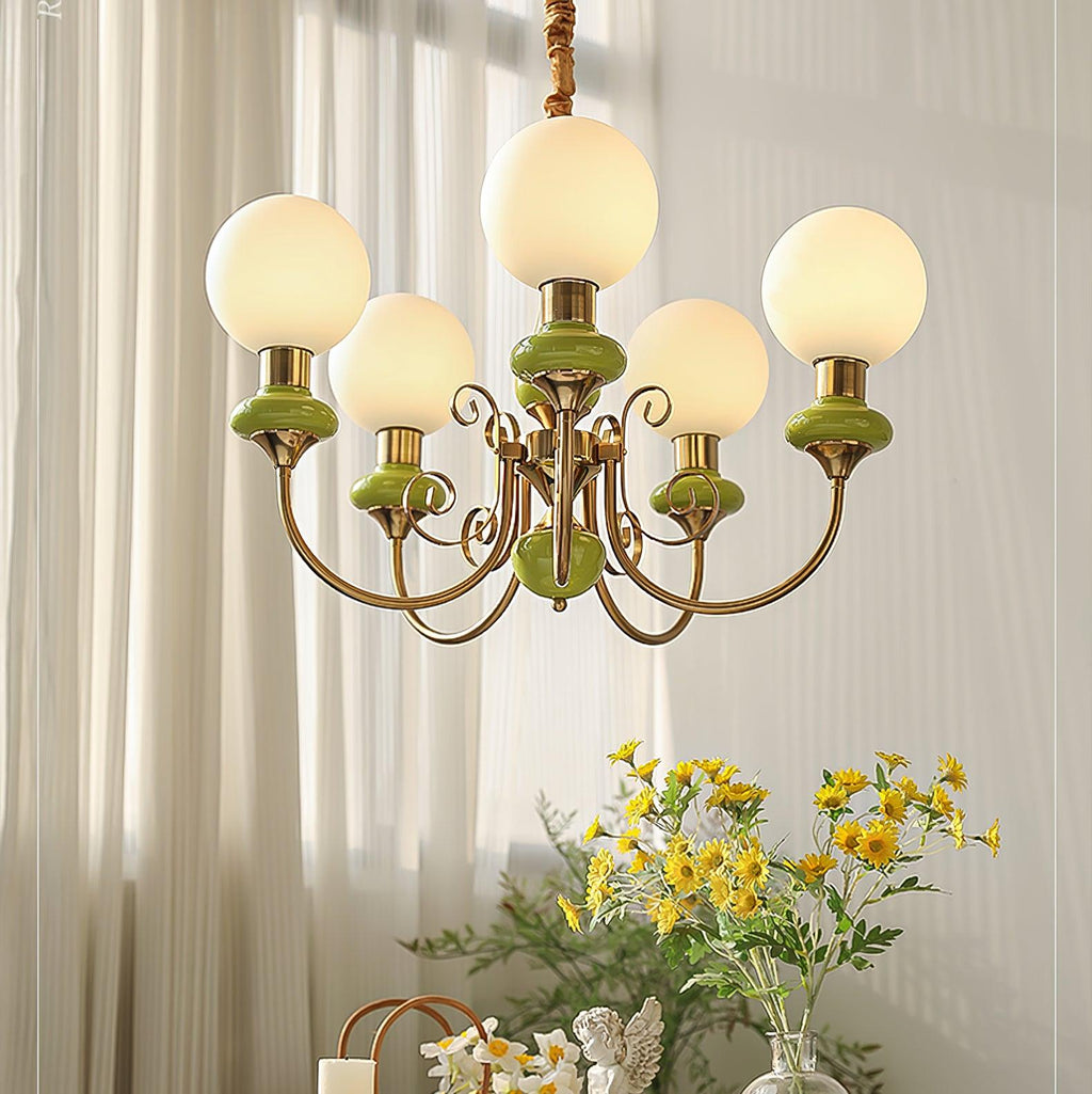 Green Clérachic Brass Scrollwork Chandelier lit against sunlit sheer curtains, yellow daisies on the table below.