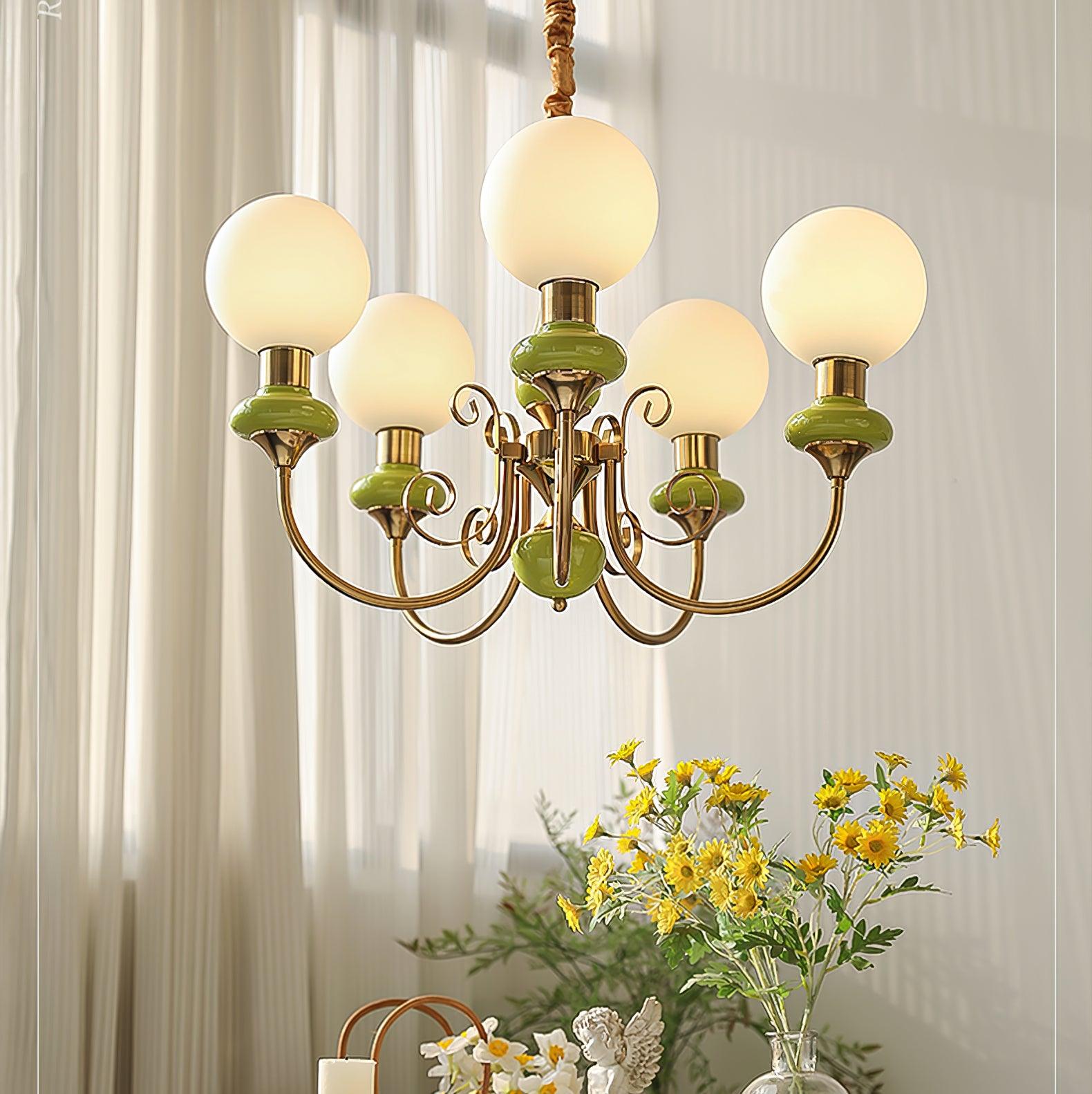 Green Clérachic Brass Scrollwork Chandelier lit against sunlit sheer curtains, yellow daisies on the table below.