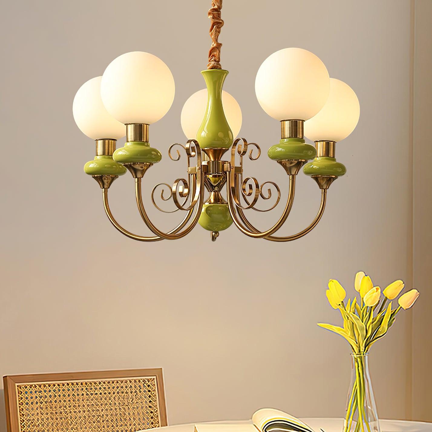 Green five-light Clérachic Brass Scrollwork Chandelier lit over a dining table set with yellow tulips and a rattan chair.