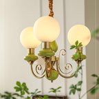 Clérachic Brass Scrollwork Chandelier in green shown close, three globes lit against a panelled wall with trailing ivy.