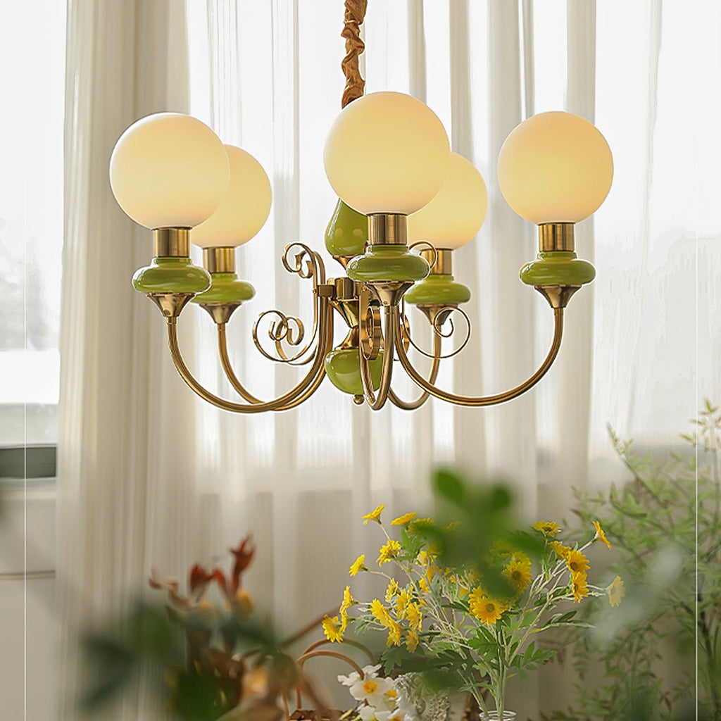 Green Clérachic Brass Scrollwork Chandelier lit in front of window sheers, spring flowers on the table below.