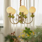 Green Clérachic Brass Scrollwork Chandelier lit in front of window sheers, spring flowers on the table below.