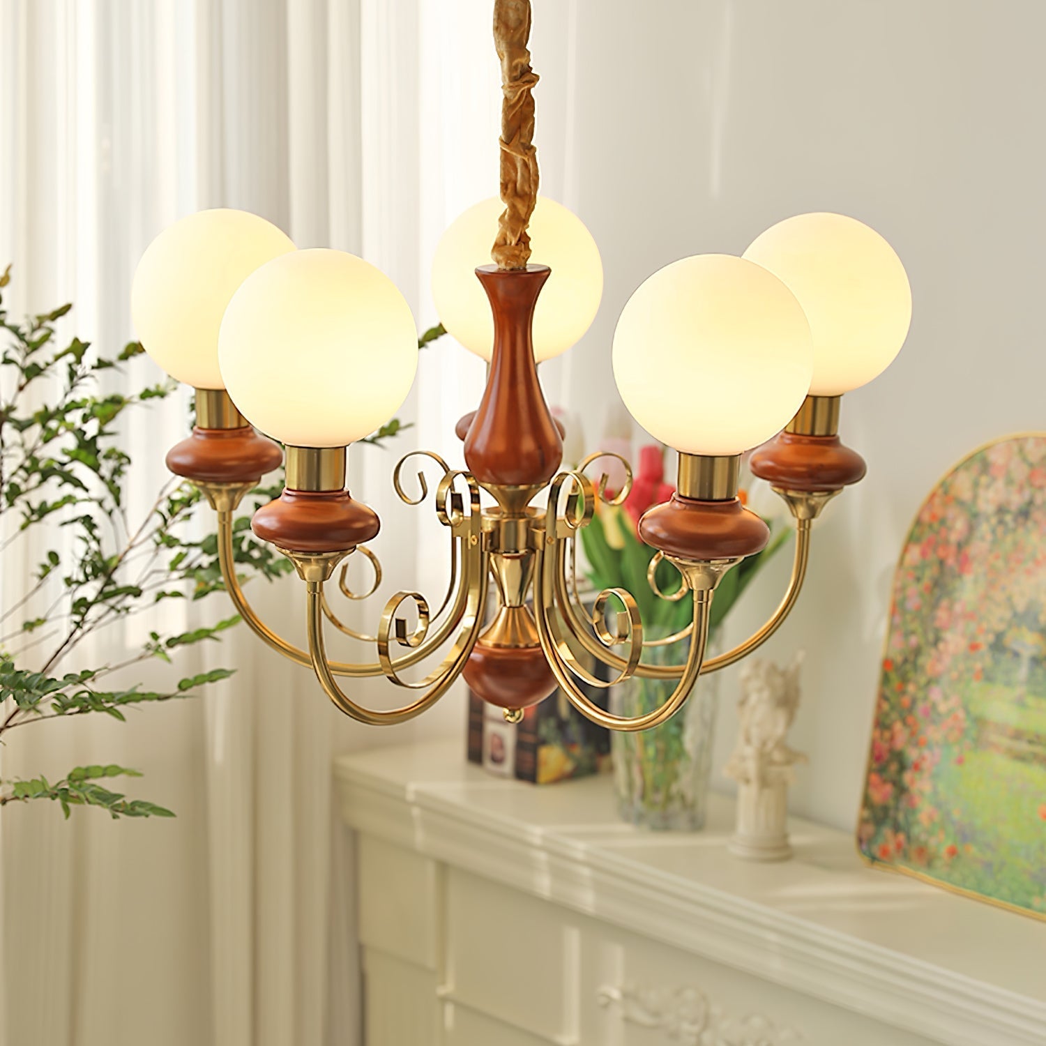 Red-brown Clérachic Brass Scrollwork Chandelier lit beside a mantel set with tulips and a framed garden painting.