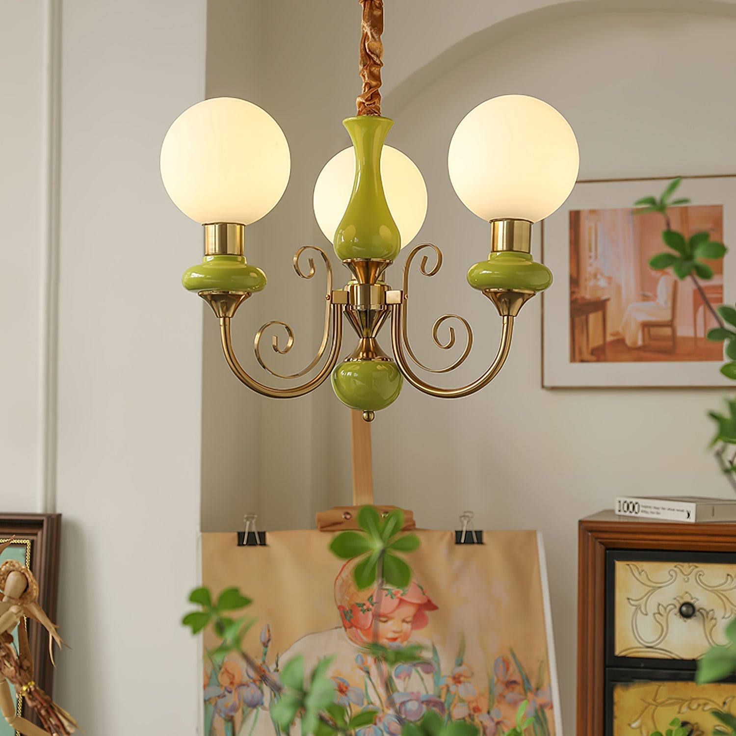 Clérachic Brass Scrollwork Chandelier in green, three lit globes above an easel in a softly arched painting corner.