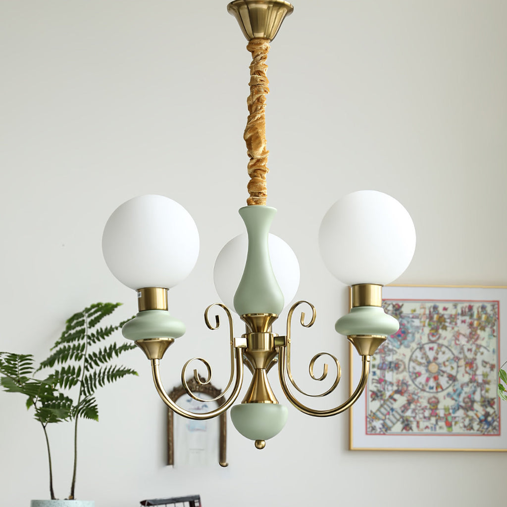 Three-light sage-green Clérachic Brass Scrollwork Chandelier lit beside a framed print and a fern in a bright corner.