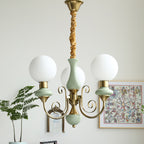 Three-light sage-green Clérachic Brass Scrollwork Chandelier lit beside a framed print and a fern in a bright corner.