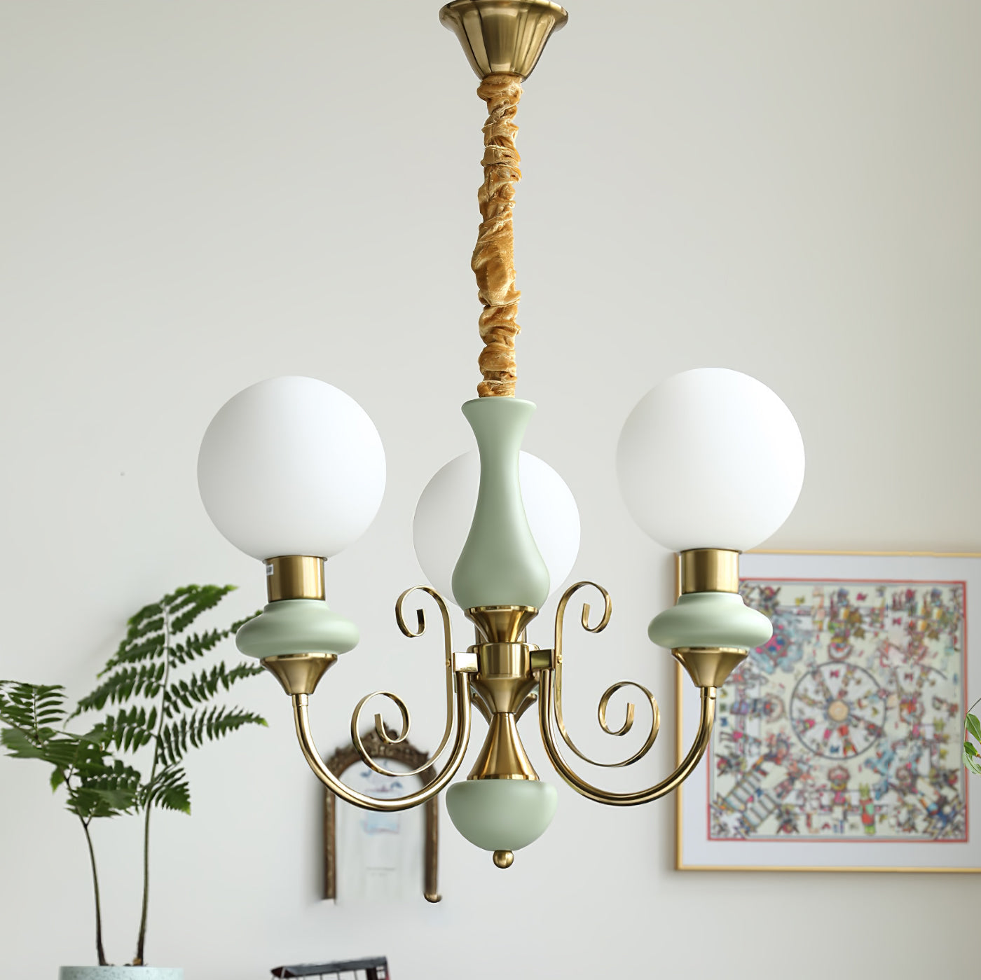 Three-light sage-green Clérachic Brass Scrollwork Chandelier lit beside a framed print and a fern in a bright corner.