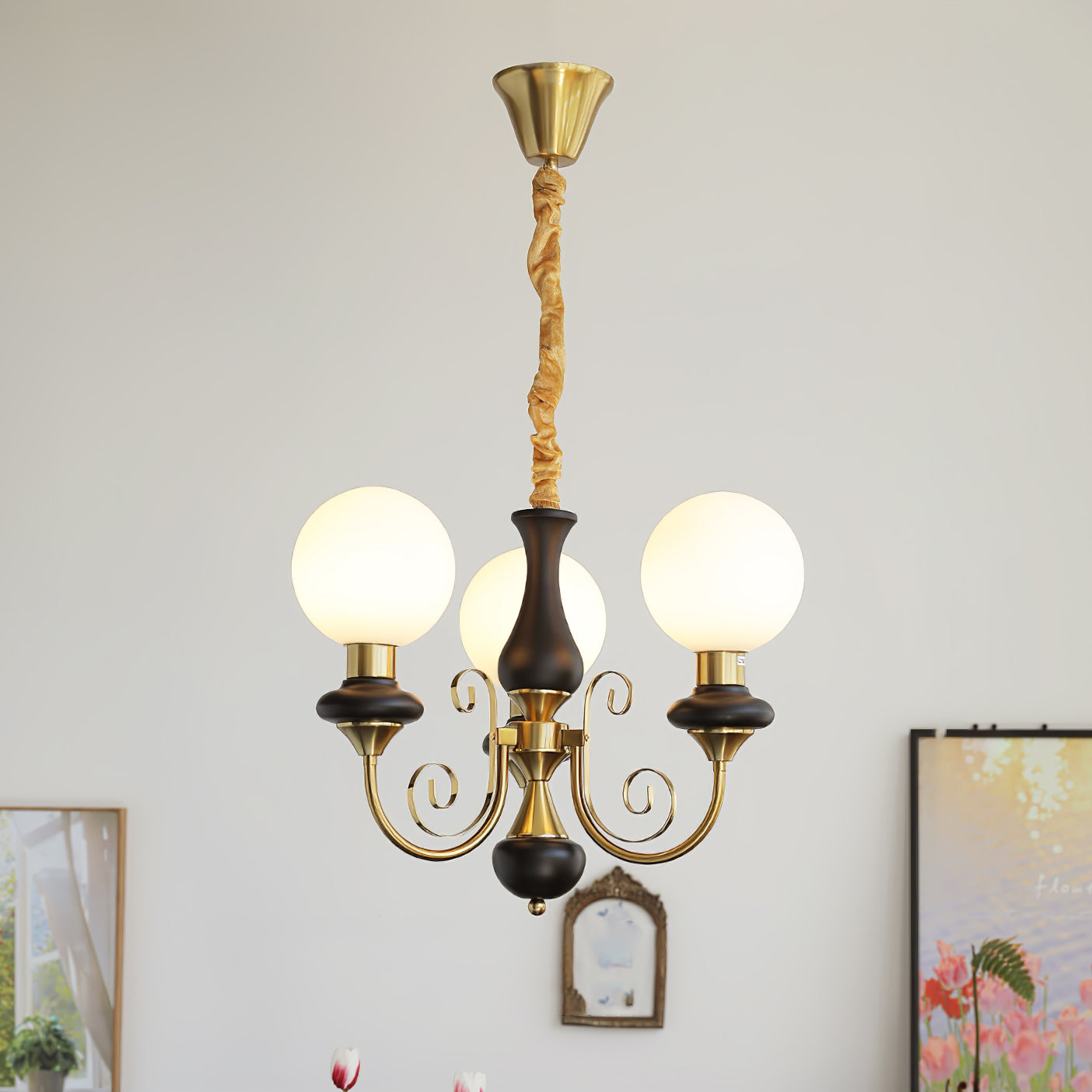 Clérachic Brass Scrollwork Chandelier in black, three lit globes on a plain wall between two framed pictures.