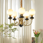 Black Clérachic Brass Scrollwork Chandelier with five globes lit in front of sunlit sheers, tulips and foliage nearby.