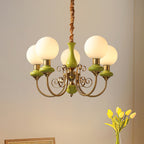 Green Clérachic Brass Scrollwork Chandelier lit against a plain warm wall, yellow tulips on the table beneath it.