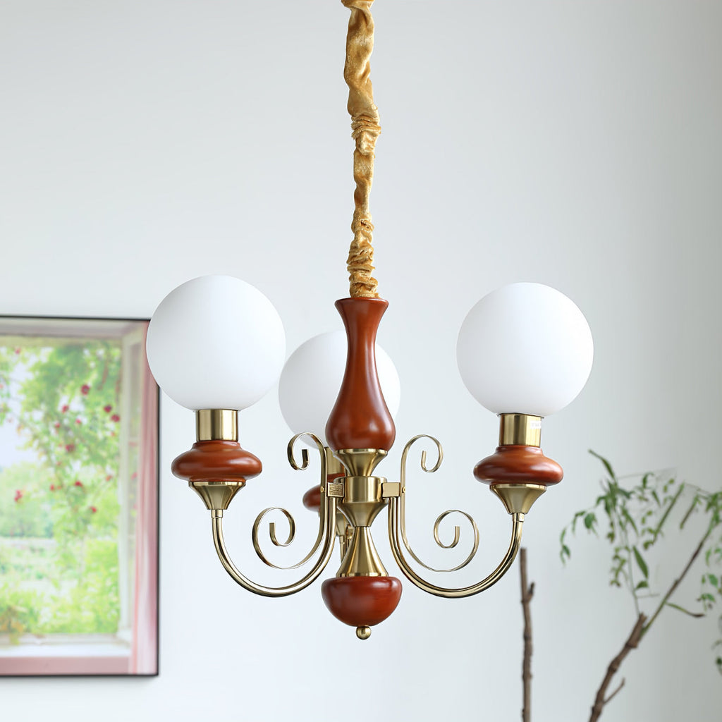Three-light Clérachic Brass Scrollwork Chandelier in red-brown, lit beside a window looking onto a flowering garden.