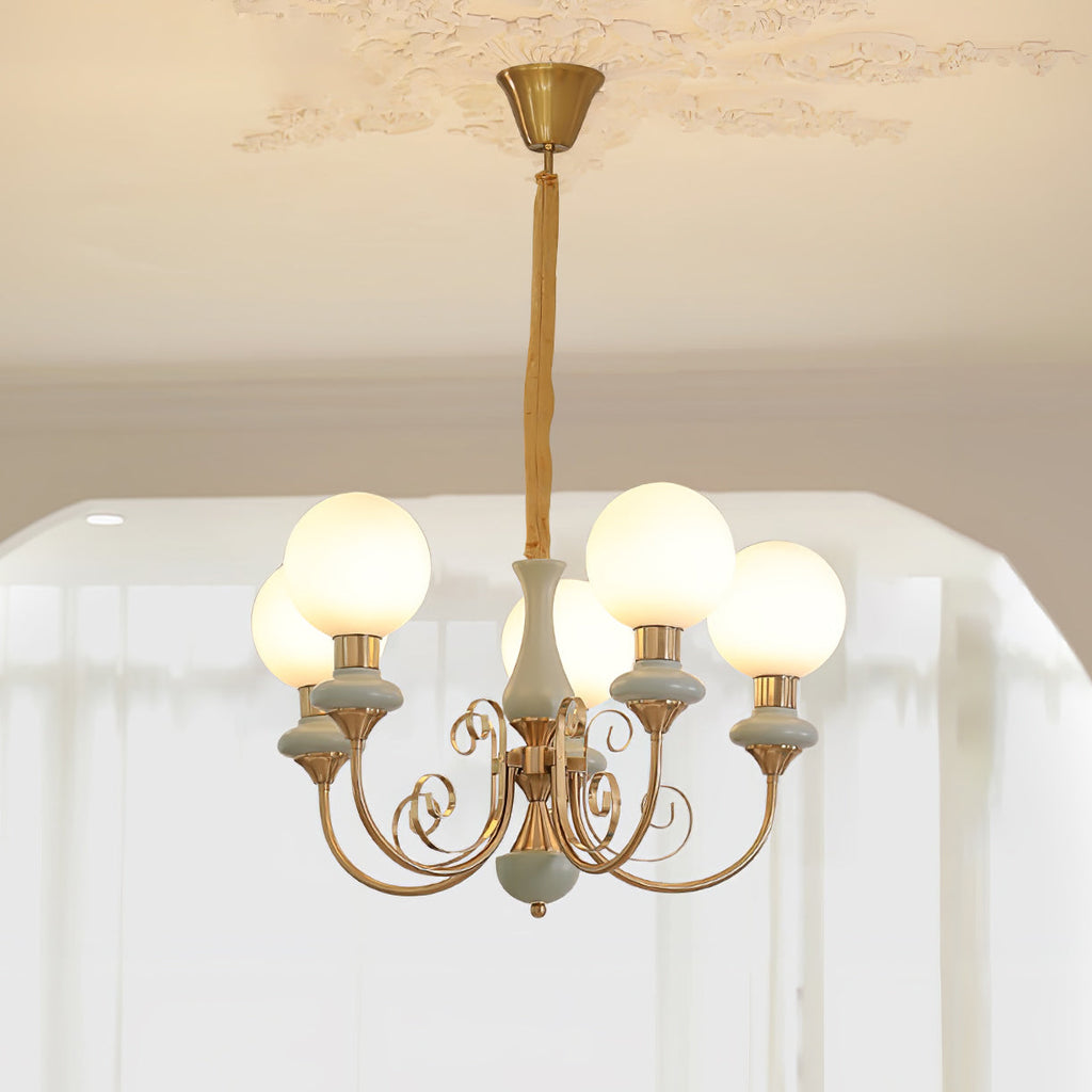 Sage-green Clérachic Brass Scrollwork Chandelier with five lit globes, hung from a moulded rose above sheer curtains.