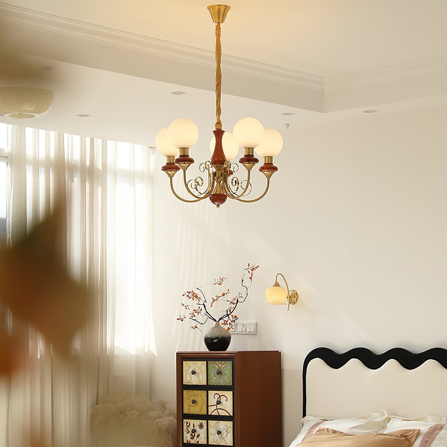 Red-brown Clérachic Brass Scrollwork Chandelier lit in a bedroom above a painted cabinet and a wavy black headboard.