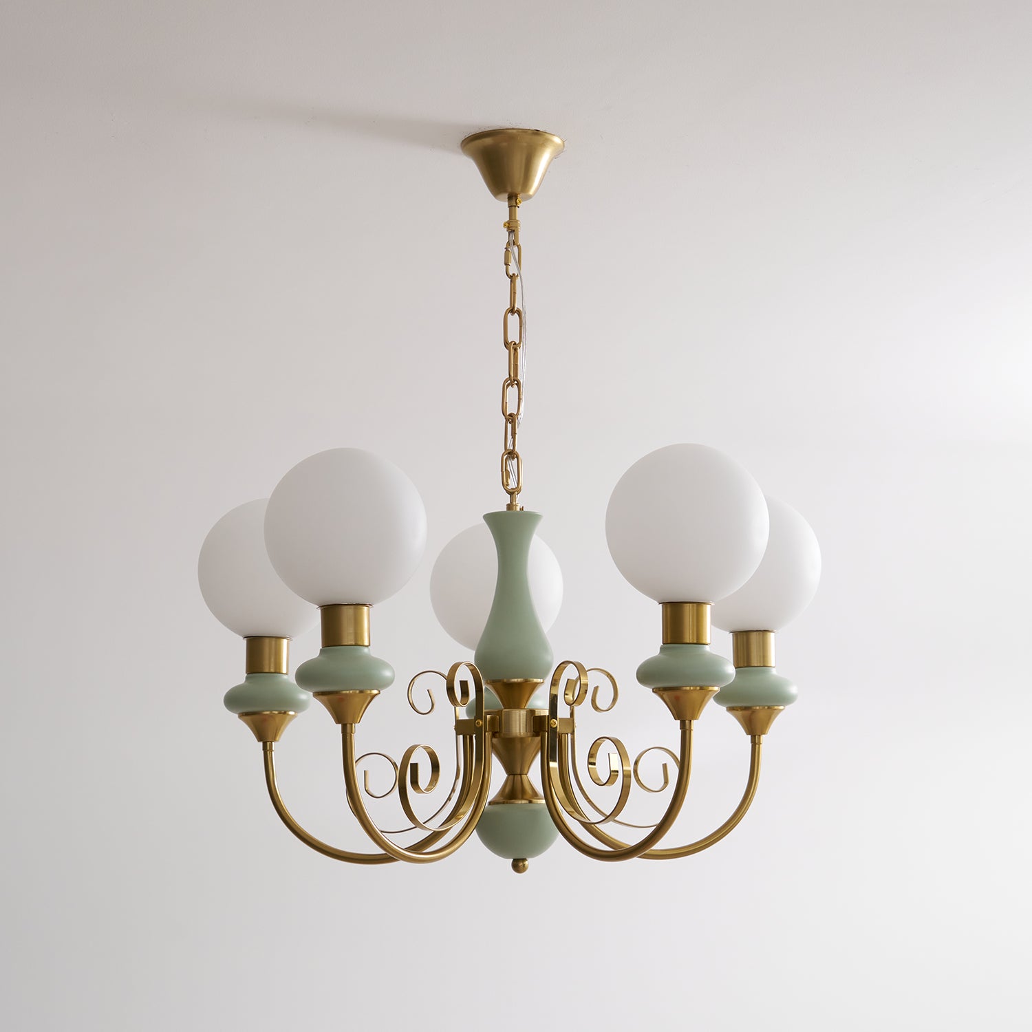 Sage-green Clérachic Brass Scrollwork Chandelier shown unlit on a brass chain against a plain white studio wall.