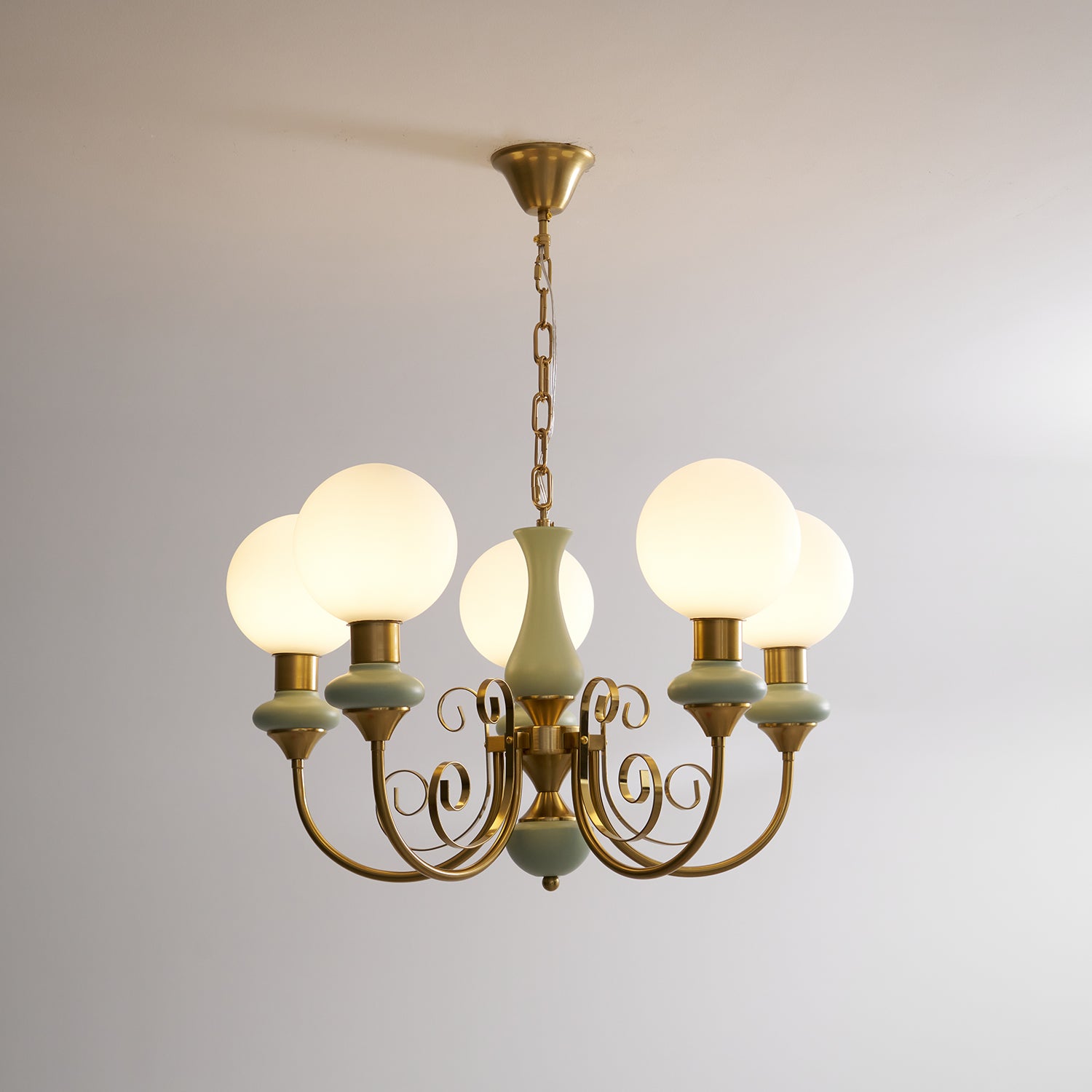 Clérachic Brass Scrollwork Chandelier in sage green on a brass chain, five opal globes lit against a bare wall.