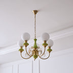 Green Clérachic Brass Scrollwork Chandelier shown unlit against a panelled wall, five matte opal globes on brass arms.