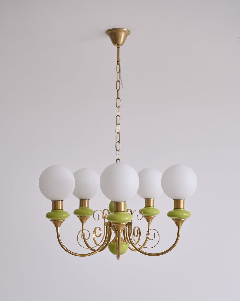Clérachic Brass Scrollwork Chandelier in green on a long brass chain, five unlit opal globes against a plain wall.