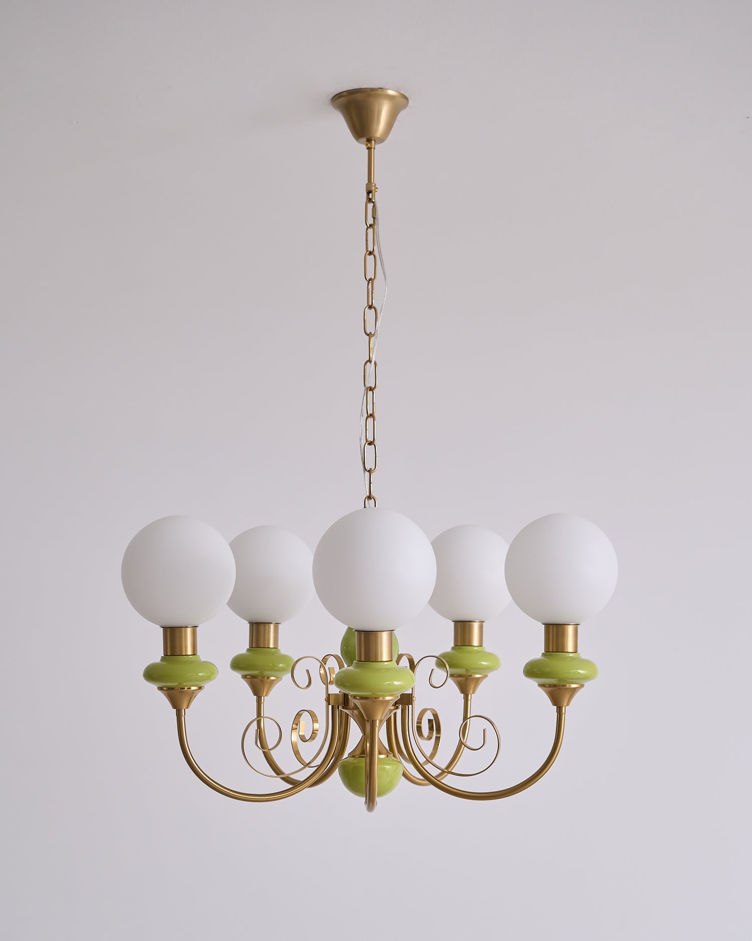 Clérachic Brass Scrollwork Chandelier in green on a long brass chain, five unlit opal globes against a plain wall.
