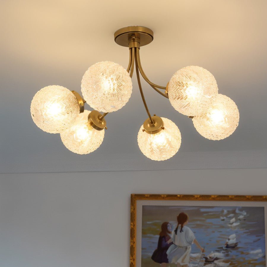 Clérachic six-head brass ceiling light glowing warm, its textured glass globes hung above a framed painting in a pale room.