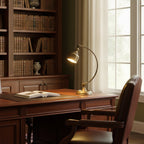 A Clérachic brass desk lamp glows on a dark wooden desk beside an open book, with a tall bookcase and window behind.