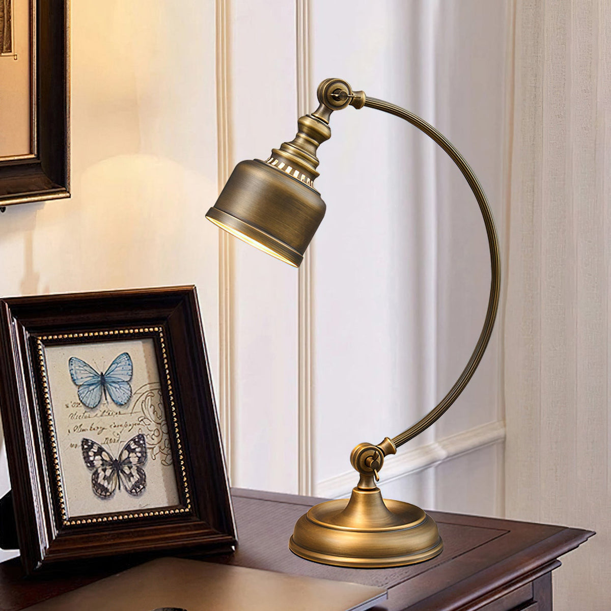 The curved brass arm of this Clérachic lamp arcs over a desk, its shade angled down towards a framed butterfly print.