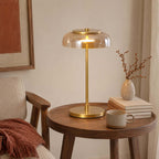 Lit Clérachic table lamp with an amber dome on a round walnut side table, beside a stack of books, a mug and a rust throw.
