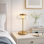 Clérachic table lamp with a clear glass dome lit on a white bedside table, beside a vase of greenery and a pile of books.
