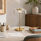 Clérachic table lamp with a clear glass dome lit on a pale work table, beside a pen pot, a wooden tray and a small vase.