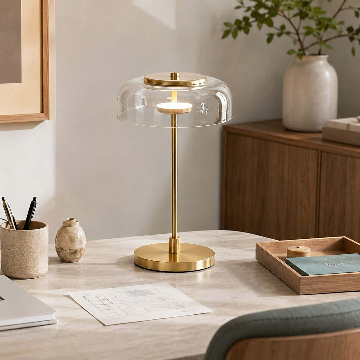 Clérachic table lamp with a clear glass dome lit on a pale work table, beside a pen pot, a wooden tray and a small vase.