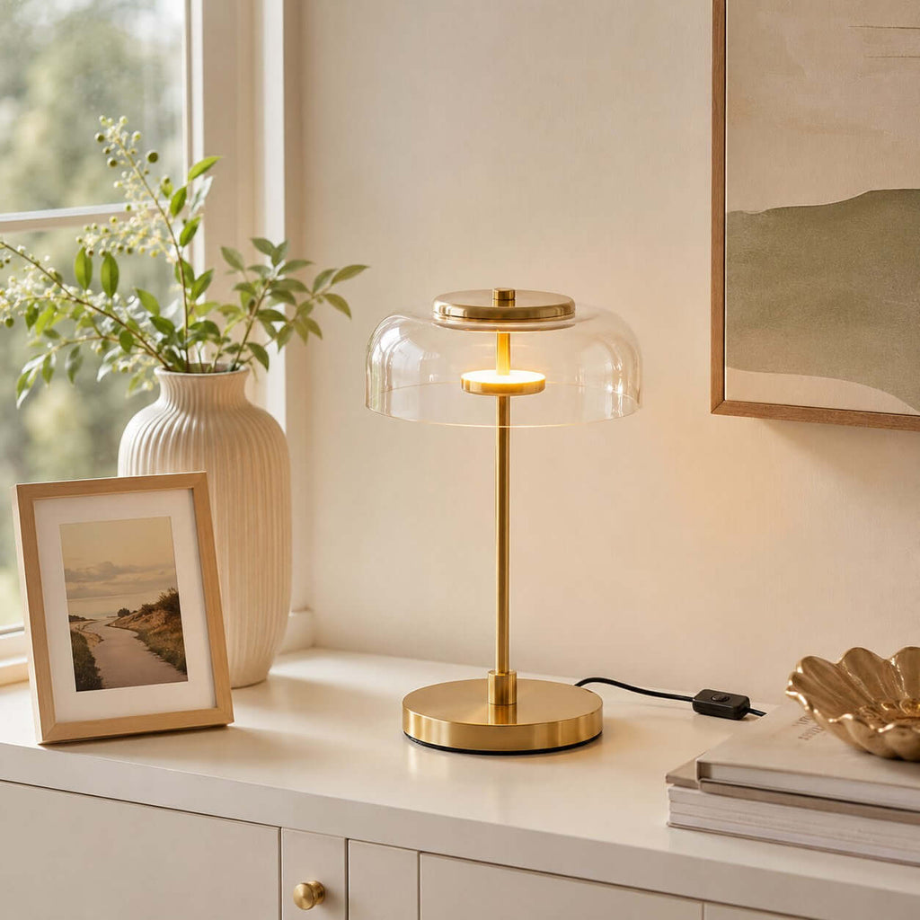 Clérachic table lamp with a clear glass dome lit on a white cabinet, beside a framed picture and a ribbed vase of leaves.