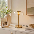 Clérachic table lamp with a clear glass dome lit on a white cabinet, beside a framed picture and a ribbed vase of leaves.