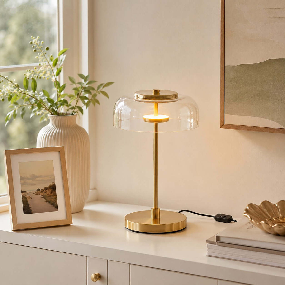 Clérachic table lamp with a clear glass dome lit on a white cabinet, beside a framed picture and a ribbed vase of leaves.