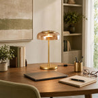 Clérachic table lamp with an amber dome on a round wooden desk, beside a laptop, a pen pot, glasses and a notebook.