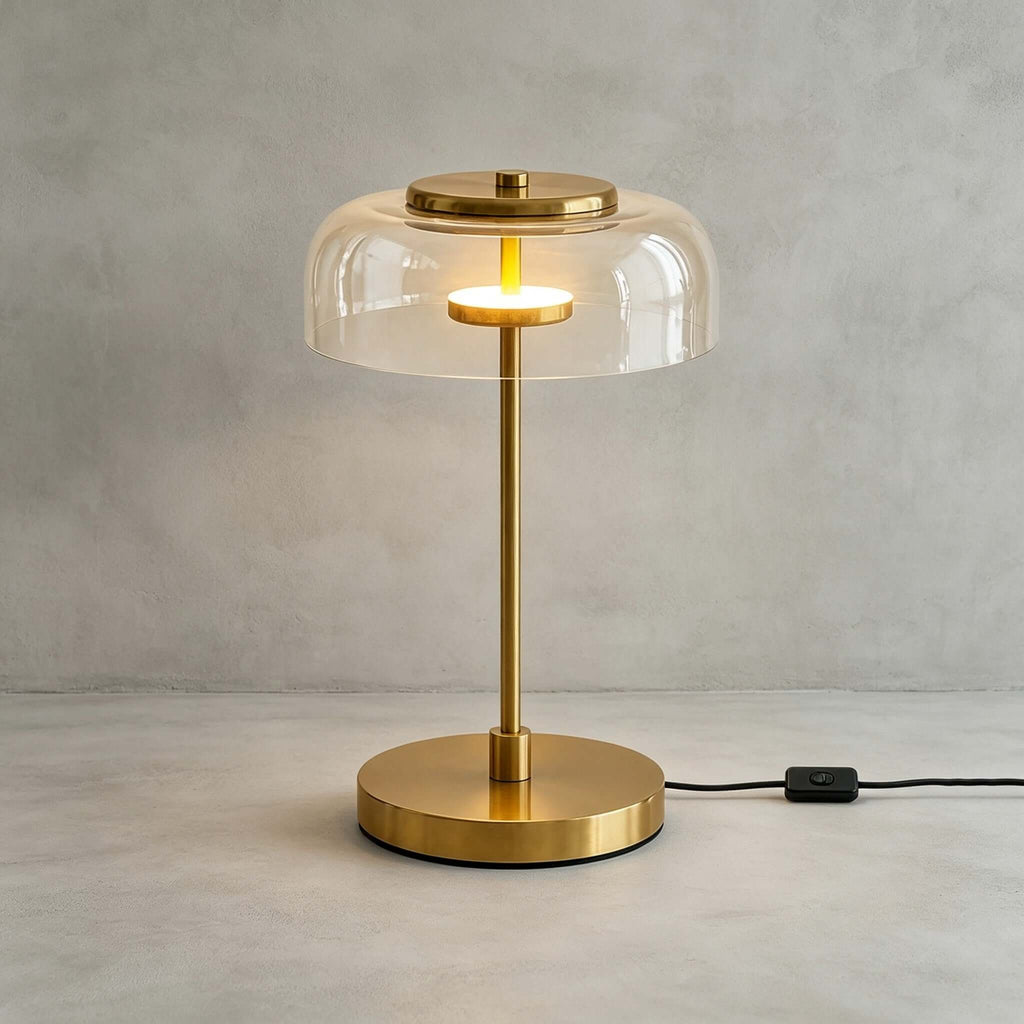 Clérachic table lamp with a clear glass dome lit against a grey wall, the brass cap and glowing ring visible under the glass.
